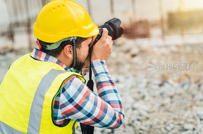 年轻男性建筑工人/工程师/建筑师/检查员/经理在建筑工地用专业相机拍摄建筑工地的照片。建筑/建筑行业概念。